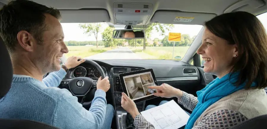 Ein Paar sitzt entspannt im Auto auf dem Weg zum K&S Farbkonzepte Showroom in Beckdorf. Die Frau zeigt dem Fahrer auf dem Tablet Bilder des aktuellen Badezimmers und hält eine handgezeichnete Grundriss-Skizze für die anstehende Renovierung bereit.