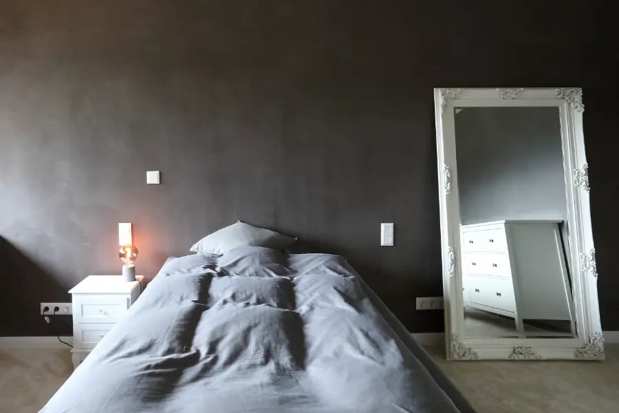 Sumpfkalkputz im Schlafzimmer in Hollenstedt: dunkelgraue, mineralische Wand, Nachttischlampe, großer weißer Standspiegel.