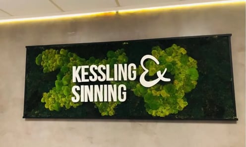 Wanddekoration mit dem Firmenlogo „Kessling &amp; Sinning“ aus weißem 3D-Schriftzug auf einer begrünten Mooswand. Das Logo befindet sich in einem schwarzen Rahmen, montiert an einer grauen Wand mit indirekter LED-Beleuchtung.