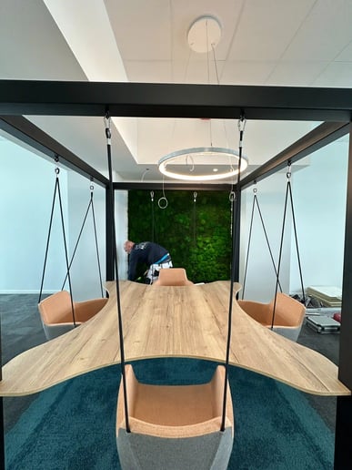 Moderner Besprechungsraum mit hängendem Holztisch und Schaukelstühlen, grünem Mooswand-Panel im Hintergrund und Frank Sinning bei der Montage.